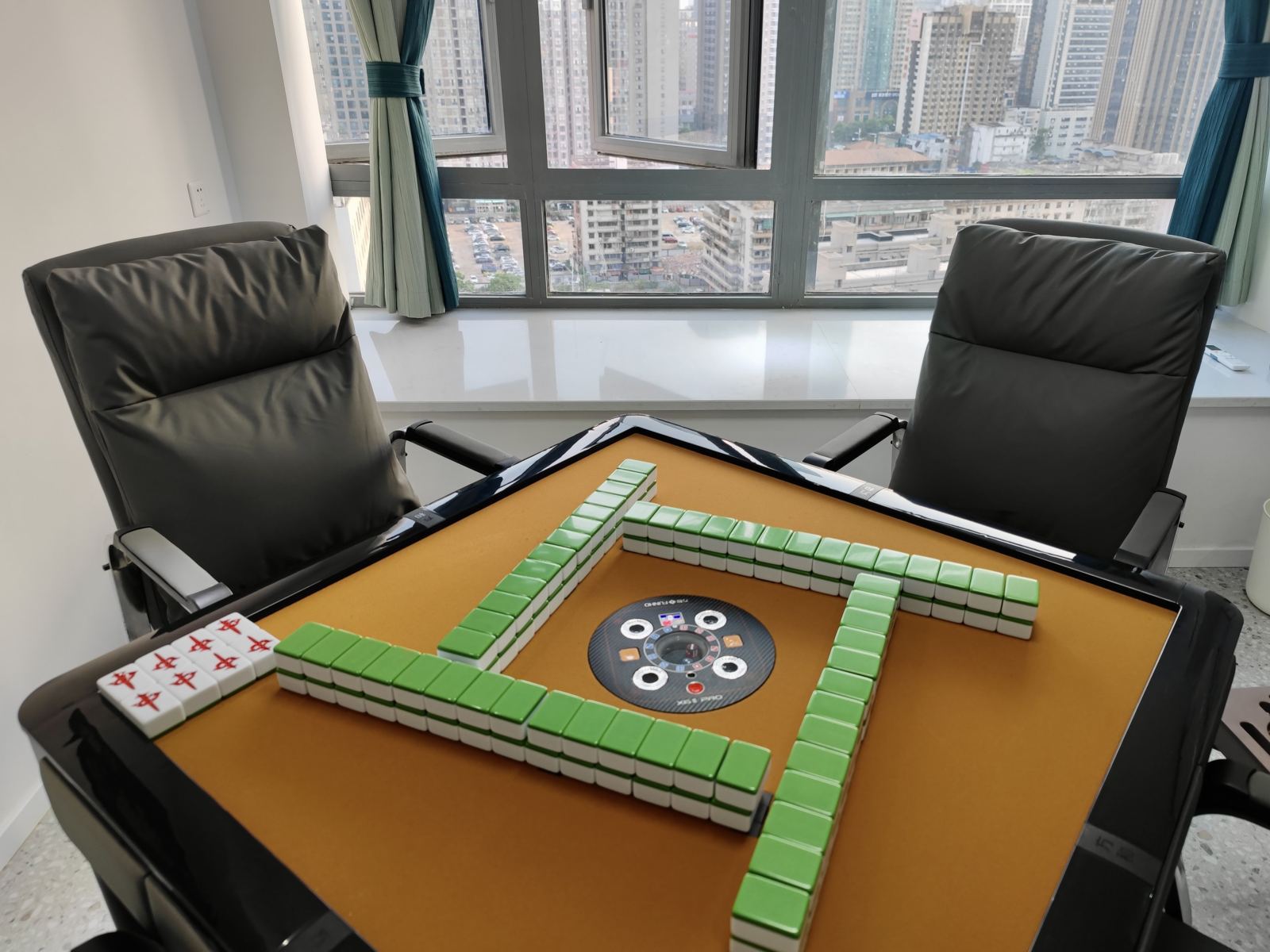 A mahjong table with green tiles laid out, city towers in the window behind